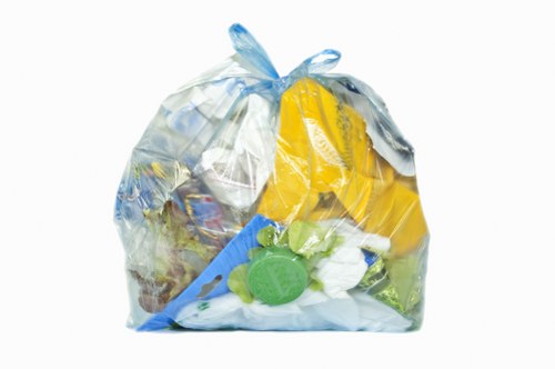 A clear plastic rubbish bag filled with mixed waste materials, including plastic bottles, packaging, and a few pieces of paper, situated on a paved surface outdoors. The bag appears semi-transparent, with a slightly crumpled texture, and is tied at the top with a knot. Within the bag, various items such as blue and yellow plastic packaging, white paper or tissue, and green plastic bottles or caps can be seen. The surrounding environment suggests an urban or residential area, possibly a driveway or pavement, with no other objects or background details visible. The image conveys the typical appearance of collected waste ready for removal, in line with rubbish collection services like those provided by Commercial Waste New Cross, serving the New Cross postcode area.