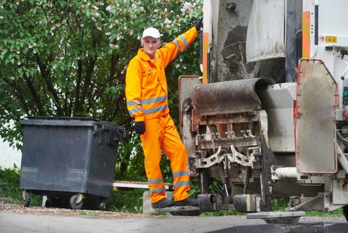 A waste collection operative dressed in high-visibility orange and reflective safety gear stands next to a large industrial rubbish compactor truck on a paved street, with one foot on the ground and the other resting on a step of the vehicle. The operative, wearing a white cap, is smiling towards the camera. Nearby on the left, there is a large black wheeled rubbish bin positioned on the sidewalk, ready for waste collection. The background features dense green foliage and trees, indicating a suburban or urban area with natural surroundings, possibly near New Cross. The scene takes place during daylight hours, with natural light illuminating the area clearly. The rubbish truck, equipped with a large cylindrical compaction mechanism, is parked with its rear section accessible for collection, emphasizing professional waste management activities carried out by Commercial Waste New Cross, a local rubbish removal service provider specializing in commercial waste disposal solutions.