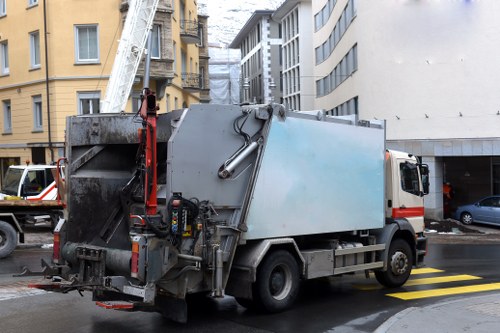 A grey and white commercial waste collection truck parked on a city street, adjacent to a building with yellow and beige facades. The truck features a large, rectangular metal waste compartment with some signs of dirt and wear, indicating frequent use. Extending from the back of the vehicle is a hydraulic arm, with a platform and lever mechanism, used for lifting and emptying waste bins. The street has a paved surface with a yellow hatch marking near the vehicle, and other parked cars are visible nearby, including a black car and a white van. Surrounding buildings include multi-story structures with balconies and windows, typical of urban environments in New Cross, London. The scene is captured during daylight, with natural lighting highlighting the industrial function of the waste collection service provided by Commercial Waste New Cross, reflecting their rubbish removal activities within the local area.